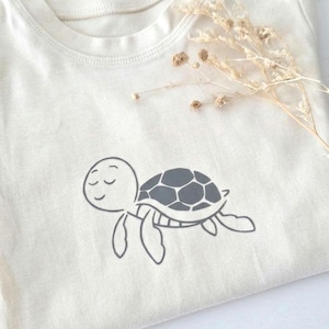 May include: A cream-coloured t-shirt featuring a dark grey graphic of a small turtle with closed eyes. The turtle has a simple, rounded shell design with visible segments. A sprig of dried, light-coloured flowers rests near the neckline.