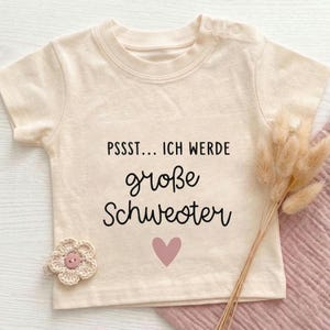 Puede incluir: Una camiseta de manga corta color crema con el texto en negro PSSST... ICH WERDE große Schwester y un pequeño corazón rosa. La prenda incluye dos botones pequeños en el cuello y un adorno de flor tejida a ganchillo.