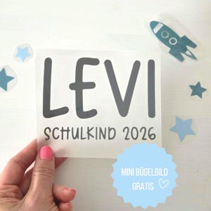 Puede incluir: Una hoja de transferencia térmica transparente con el nombre LEVI en letras grandes de color gris oscuro y el texto SCHULKIND 2026 debajo. La hoja está rodeada de motivos térmicos separados, incluyendo un cohete azul cerceta y estrellas azul claro.