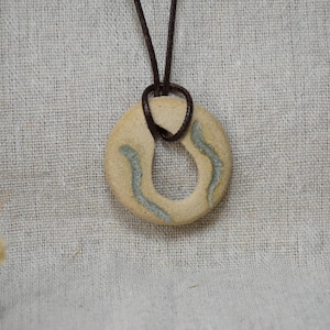 Pode incluir: Um pendente circular de cerâmica bege com duas linhas decorativas curvas em azul-acinzentado claro. O pendente está suspenso por um cordão castanho escuro que passa pela abertura central, criando um design de colar simples e funcional.