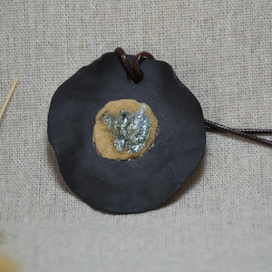 Può includere: Un ciondolo grigio scuro con un cordino in pelle marrone. Il ciondolo ha una forma circolare con un centro beige chiaro e un disegno color argento al centro. Il ciondolo è su uno sfondo di tessuto color neutro.