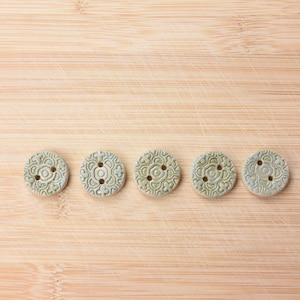 Peut inclure: Cinq boutons ronds en céramique vert clair, ornés de motifs floraux et percés de trois trous, disposés sur une surface en bois brun clair. Les boutons ont un aspect légèrement texturé. Chaque bouton mesure environ 2,5 cm de diamètre.