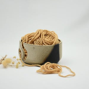Op de afbeelding: Een keramische garenkom met een bruin en zwart ontwerp, gevuld met bruin garen. De kom heeft een gebogen sleuf waar het garen doorheen kan. Een klein takje gedroogde bloemen ligt links van de kom, met een streng garen ervoor.