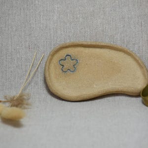 Op de afbeelding: Een beige, onregelmatig niervormig keramieken schaaltje met een kleine, donkerblauwe, vijfbladige bloemomtrek op het oppervlak. Naast het schaaltje liggen twee goudkleurige metalen vingerhoeden en een klein bosje gedroogd beige gras.