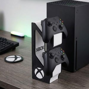 Pode incluir: Um suporte vertical para comandos de jogo em preto e branco com o logótipo e o texto Xbox. O suporte segura dois comandos sem fios pretos, um sobre o outro, com uma consola de jogos preta posicionada verticalmente ao lado da unidade.