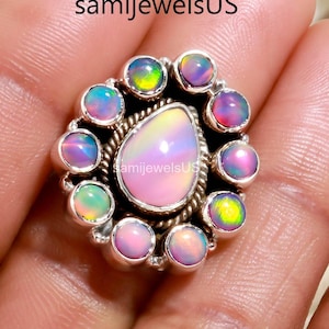 Op de afbeelding: Een zilveren ring met een peervormige opaal in het midden, omringd door een halo van kleinere opalen. De opalen vertonen een scala aan kleuren, waaronder roze, groen en blauw. De ring wordt in een hand gehouden.