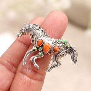 May include: A silver-colored horse figurine with intricate detailing. The horse is adorned with orange and green gemstones, adding a pop of color. The figurine is a decorative item, possibly a brooch or pendant.
