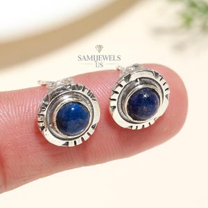 Könnte beinhalten: Ein Paar silberne Ohrstecker mit einem runden, dunkelblauen Edelstein in der Mitte. Die Ohrringe haben ein strukturiertes, kreisförmiges Design um den Edelstein herum. Der Markenname "SAMIJEWELS US" ist sichtbar.