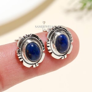 May include: Silver stud earrings featuring oval, deep blue gemstones. Each earring has a decorative, textured border. The image is well-lit, highlighting the jewellery's details. The earrings are displayed on a light background.