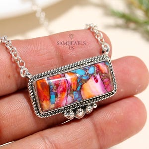 May include: A silver necklace featuring a rectangular pendant with a colorful, patterned stone. The stone displays shades of pink, orange, blue, and brown. The pendant is framed with a twisted rope design and small decorative accents.