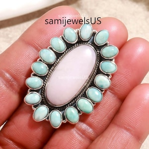 May include: Silver ring with an oval light pink central stone, encircled by small, oval, light blue-green stones. The ring is held in a hand. The text "samijewelsUS" is visible.