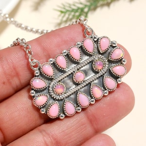 Puede incluir: Un collar de plata con un colgante rectangular con piedras preciosas de ópalo rosa. El colgante tiene un diseño detallado con una fila central de ópalos más pequeños y un borde de ópalos más grandes en forma de lágrima. La cadena es delicada y plateada.