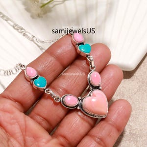 May include: A silver chain necklace featuring alternating pink and turquoise heart-shaped and oval stones set in metal bezels. The design includes a large central heart-shaped pink stone and the text samijewelsUS printed on the image.