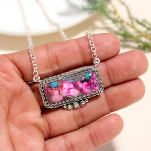 May include: A silver necklace featuring a rectangular pendant with a vibrant, multi-colored stone. The stone displays shades of pink, purple, and turquoise, set within a decorative silver frame. The necklace is held in a hand, showcasing the intricate design.