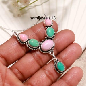 May include: A silver necklace featuring alternating oval and teardrop-shaped pink and green stones set in decorative metal frames. The piece includes a central pendant drop and the text samijewelsUS is visible on the metal surface of the jewelry.