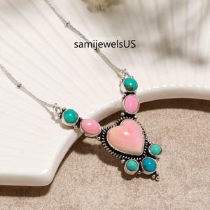 Puede incluir: Un collar de plata con una piedra central rosa en forma de coraz&oacute;n, acentuado con piedras redondas m&aacute;s peque&ntilde;as de color rosa y turquesa en un engaste de metal decorativo. La cadena es fina y met&aacute;lica, el texto samijewelsUS es visible.