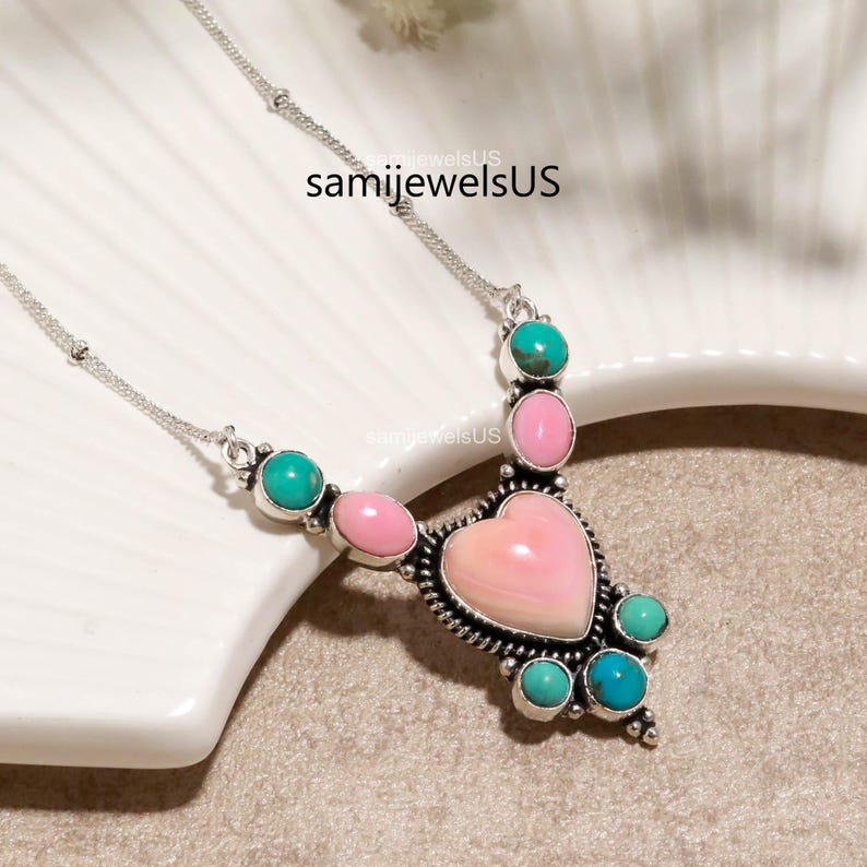 Puede incluir: Un collar de plata con una piedra central rosa en forma de coraz&oacute;n, acentuado con piedras redondas m&aacute;s peque&ntilde;as de color rosa y turquesa en un engaste de metal decorativo. La cadena es fina y met&aacute;lica, el texto samijewelsUS es visible.