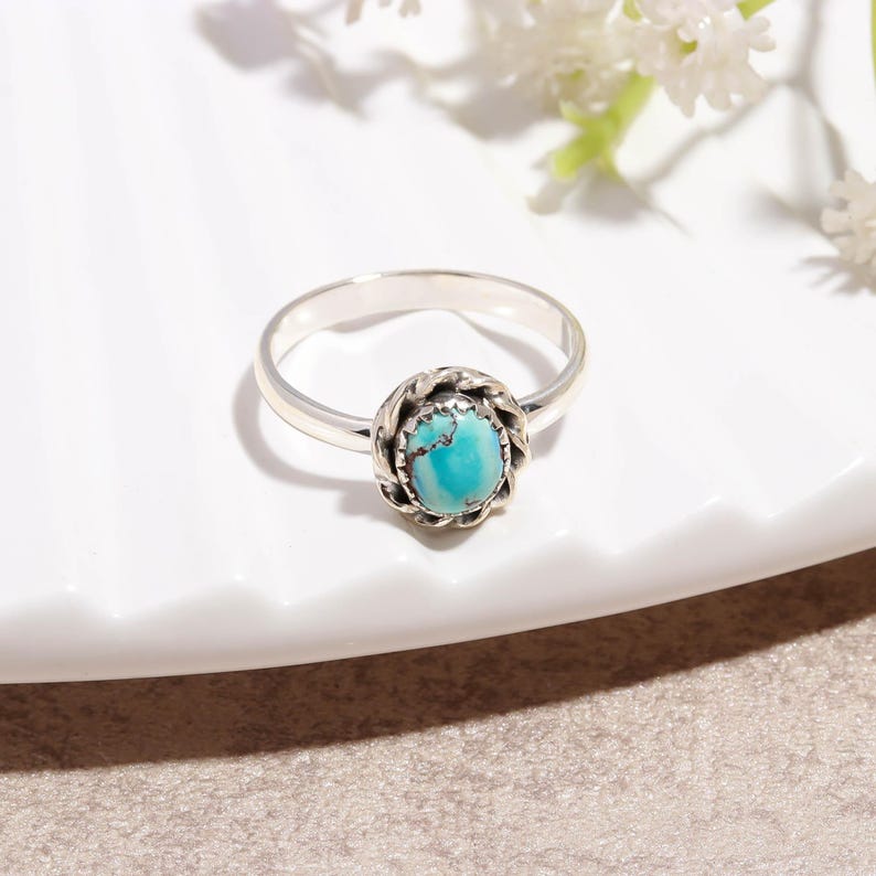 Puede incluir: Un anillo de plata con una piedra turquesa. El anillo presenta una piedra turquesa redonda de color azul claro engastada en un engaste de metal retorcido decorativo. La banda es de plata pulida simple.