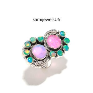 May include: A silver ring featuring two large oval pink opals and several smaller green opals. The ring has a detailed, ornate design with a floral or leaf-like pattern around the stones. The text "samijewelsUS" is visible.