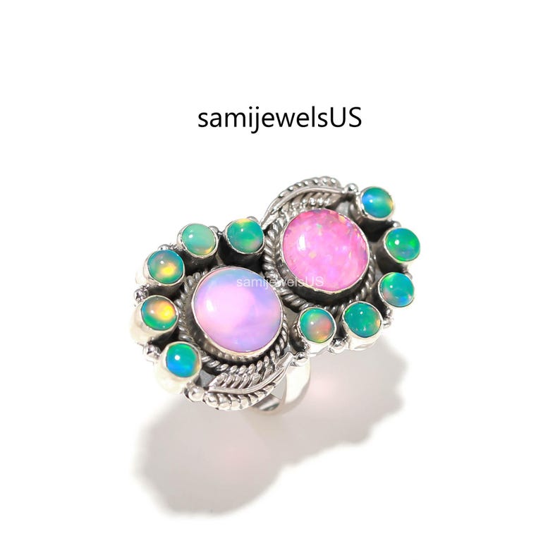 May include: A silver ring featuring two large oval pink opals and several smaller green opals. The ring has a detailed, ornate design with a floral or leaf-like pattern around the stones. The text "samijewelsUS" is visible.