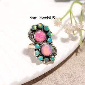 May include: A silver-toned ring featuring two large, pink opal stones surrounded by smaller, teal and green opal stones. The ring has a detailed, ornate design with leaf accents. The text "samijewelsUS" is visible.