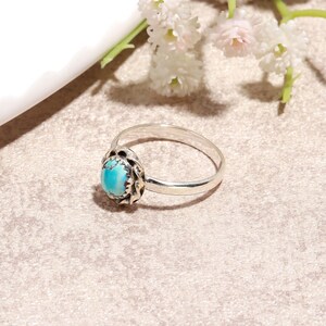 Puede incluir: Un anillo de plata con una piedra ovalada turquesa. La piedra est&aacute; engastada en un bisel decorativo. El anillo est&aacute; sobre una superficie de tonos neutros, con flores blancas en el fondo.
