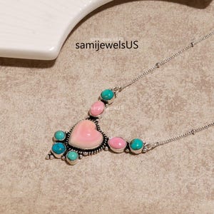 Puede incluir: Un collar de plata con una piedra central rosa en forma de coraz&oacute;n rodeada de piedras redondas m&aacute;s peque&ntilde;as de color turquesa y rosa en un engaste de metal decorativo. El texto samijewelsUS es visible sobre el colgante en la cadena.