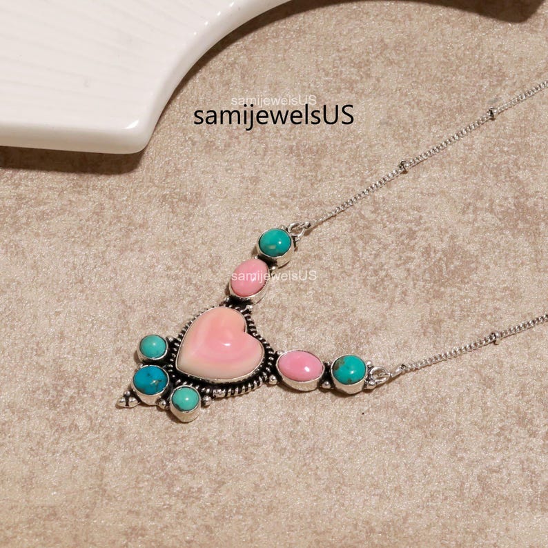 Puede incluir: Un collar de plata con una piedra central rosa en forma de coraz&oacute;n rodeada de piedras redondas m&aacute;s peque&ntilde;as de color turquesa y rosa en un engaste de metal decorativo. El texto samijewelsUS es visible sobre el colgante en la cadena.