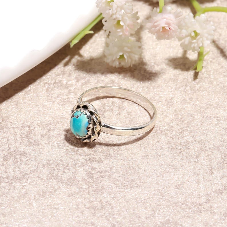 Puede incluir: Un anillo de plata con una piedra ovalada turquesa. La piedra est&aacute; engastada en un bisel decorativo. El anillo est&aacute; sobre una superficie de tono neutro, con flores blancas en el fondo.