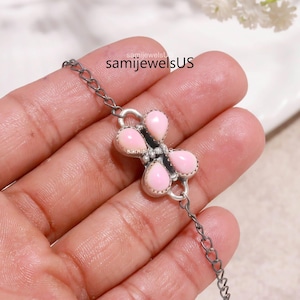 May include: A silver-toned chain bracelet featuring a central decorative element with four teardrop-shaped, smooth, light pink stones arranged in a symmetrical pattern. The metal setting has a dark, oxidised finish. The text samijewelsUS is visible.