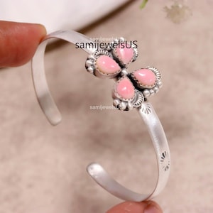 May include: A silver cuff bracelet featuring a central butterfly-shaped ornament adorned with four teardrop-shaped pink stones. The metal band displays etched sunburst patterns along its length. The text samijewelsUS is visible on the image.