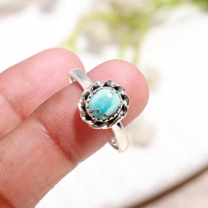 Puede incluir: Un anillo de plata con una piedra preciosa azul claro engastada en un dise&ntilde;o floral. La piedra preciosa redonda est&aacute; rodeada por un engaste de plata texturizado y detallado. La banda del anillo es lisa, creando un contraste con el engaste ornamentado.