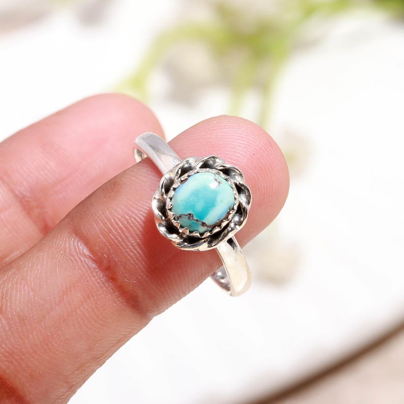 Puede incluir: Un anillo de plata con una piedra preciosa azul claro engastada en un dise&ntilde;o floral. La piedra preciosa redonda est&aacute; rodeada por un engaste de plata texturizado y detallado. La banda del anillo es lisa, creando un contraste con el engaste ornamentado.