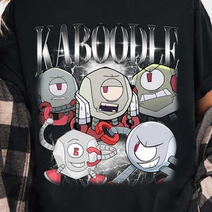Può includere: T-shirt nera con la scritta "KABOODLE" in argento sopra una grafica di cinque personaggi robot. I personaggi hanno accenti rossi e grigi. La t-shirt è indossata con una camicia a quadri nera e grigia.