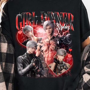 Può includere: T-shirt nero con un design grafico rosso con la scritta "GIRL DINNER Sylas". Il design include più immagini di un personaggio con i capelli argentati, su uno sfondo a forma di cuore rosso. La maglietta ha maniche corte.