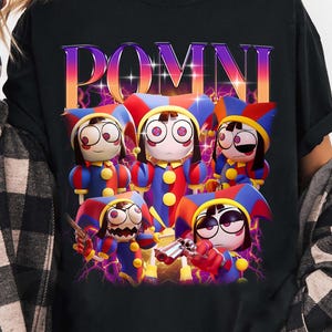 Könnte beinhalten: Schwarzes T-Shirt mit dem Wort "POMNI" in einem farbenfrohen Retro-Schriftzug. Darunter befinden sich Cartoon-Charaktere in Narrenkostümen mit roten, blauen und gelben Akzenten. Die Charaktere haben große Augen und sind von lila Blitzen umgeben.