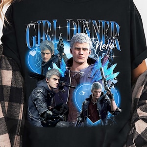 Peut inclure: T-shirt noir avec un motif graphique. Le motif comprend le texte "GIRL DINNER Nero" et plusieurs images d'un personnage aux cheveux blancs et aux accents bleus. Le t-shirt est à col rond.