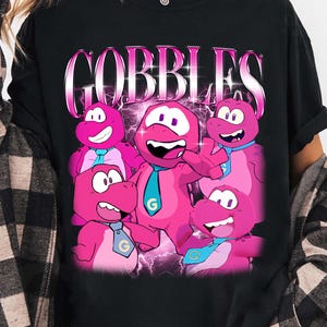 Può includere: T-shirt nera con una grafica di cinque personaggi dei cartoni animati rosa e la scritta "GOBBLES" in un font retrò. I personaggi hanno grandi occhi e indossano cravatte con la lettera "G".