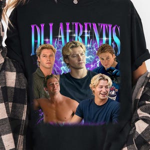 Peut inclure: Un t-shirt noir avec une impression graphique représentant plusieurs images d'un homme blond. Le texte "DILAURENTIS" est imprimé dans une grande police stylisée en dégradé bleu et violet, sur un fond d'éclairs violets.