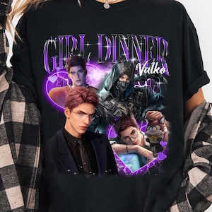 Può includere: Una t-shirt nera a maniche corte con una stampa grafica che riporta il testo "GIRL DINNER" e "Valko" in lettere viola. Il design include tre figure maschili illustrate, una delle quali indossa una maschera, e un piccolo personaggio felino tra cuori viola.
