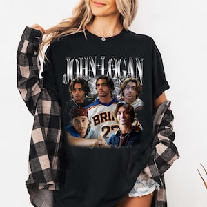 May include: A black short-sleeve t-shirt featuring the text JOHN LOGAN in a metallic font at the top. Below the text are five photographic portraits of the same person in different outfits, including a sports jersey with the number 22.