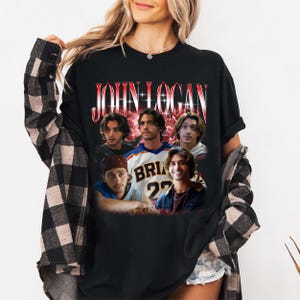 May include: A black t-shirt featuring a graphic print of the name JOHN LOGAN in red and white lettering above a collage of five photographs of the same individual in various outfits, including a sports jersey with the text BRIAN 22.