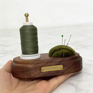以下が含まれることがあります： オリーブグリーンの糸巻きとベルベットの針刺しが付いた、手作りの木製裁縫アクセサリー。濃い茶色のベースには「HAND MADE」と書かれた真鍮プレートが付いています。クッションにはいくつかのピンが挿入されています。