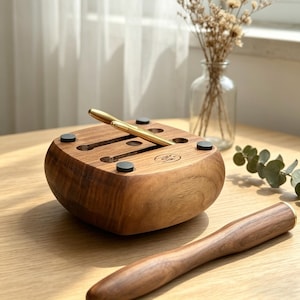 May include: A wooden pen holder with a curved design, featuring slots for pens and small, round, black pads on the base. A gold pen rests in one of the slots. A matching wooden pen is in the foreground. The holder is made of a warm-toned wood.