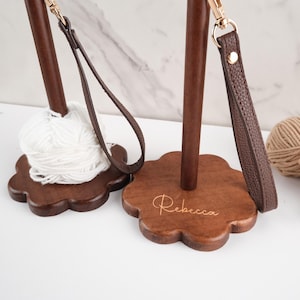 以下が含まれることがあります： 濃い茶色の仕上げが施された2つの木製糸ホルダー。1つは白い糸玉を保持し、もう1つはベースに「Rebecca」という名前が刻まれています。それぞれに、金色の留め金が付いた茶色の革ストラップが付いています。