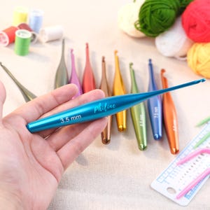 以下が含まれることがあります： 様々な色の編み棒のコレクション。焦点は3.5mmのターコイズ色の編み棒にあり、「Philine」と書かれています。他の編み棒は、金、緑、オレンジなど、さまざまな色があります。背景には糸やその他の工芸品があります。