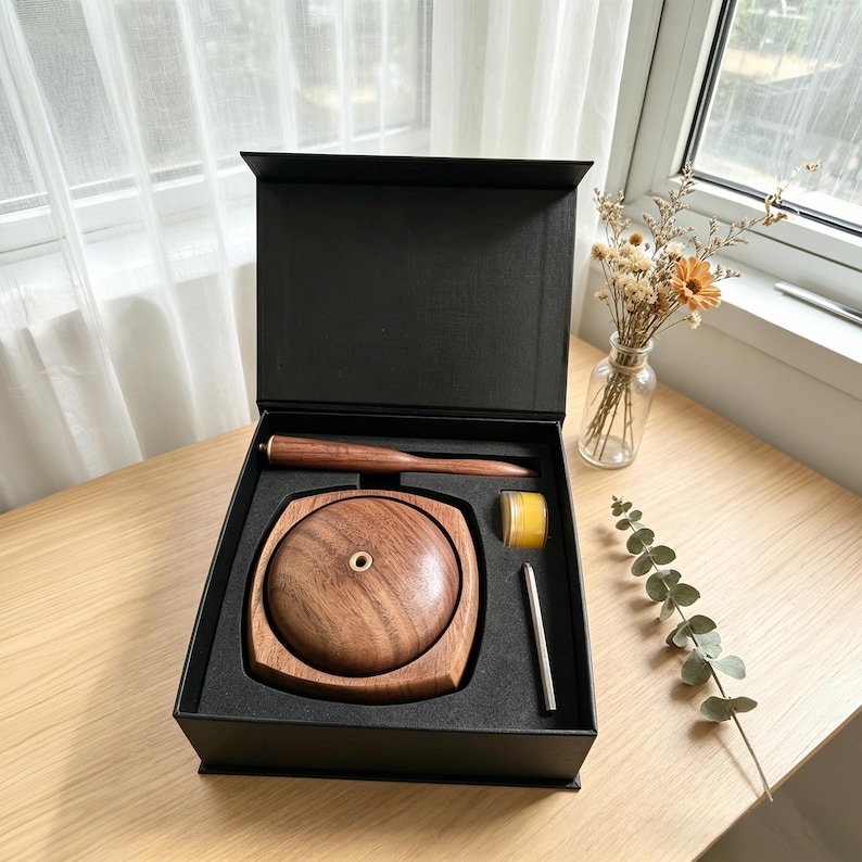 May include: A wooden incense burner set in a black box. The set includes a round wooden burner, a wooden tool, a small jar of yellow wax, and a silver metal stick. The box is open, revealing the contents.