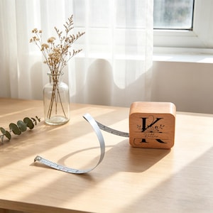 Puede incluir: Una cinta métrica de madera en forma de cubo con la letra "K" y el nombre "Katter" grabados. La cinta es plateada y se extiende desde el cubo de madera. Un jarrón de cristal transparente con flores secas está cerca.