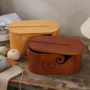 以下が含まれることがあります： 楕円形の蓋が付いた2つの木製糸収納ボックス。1つは明るい色、もう1つは濃い茶色です。濃い茶色のボックスには装飾的な渦巻き模様と「wilson」という名前が「EST 2023」とともに印刷されています。糸と編み針が見えます。