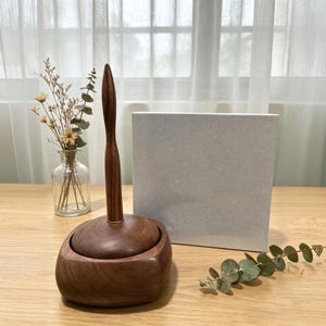 May include: A handcrafted wooden toilet paper holder with a matching lid. The holder has a dark brown finish and a tall, slender handle. A small glass vase with dried flowers and a silver box are in the background.